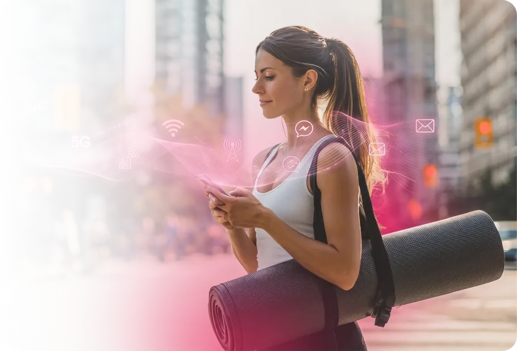 Joven mujer en ropa deportiva llevando una esterilla de yoga enrollada caminando por una calle urbana, mirando su smartphone, con íconos digitales rosas brillantes (señal Wi-Fi, burbuja de mensaje, sobre de correo electrónico) flotando a su alrededor, que representa a una usuaria móvil recibiendo comunicaciones multicanal dirigidas de un operador de telecomunicaciones