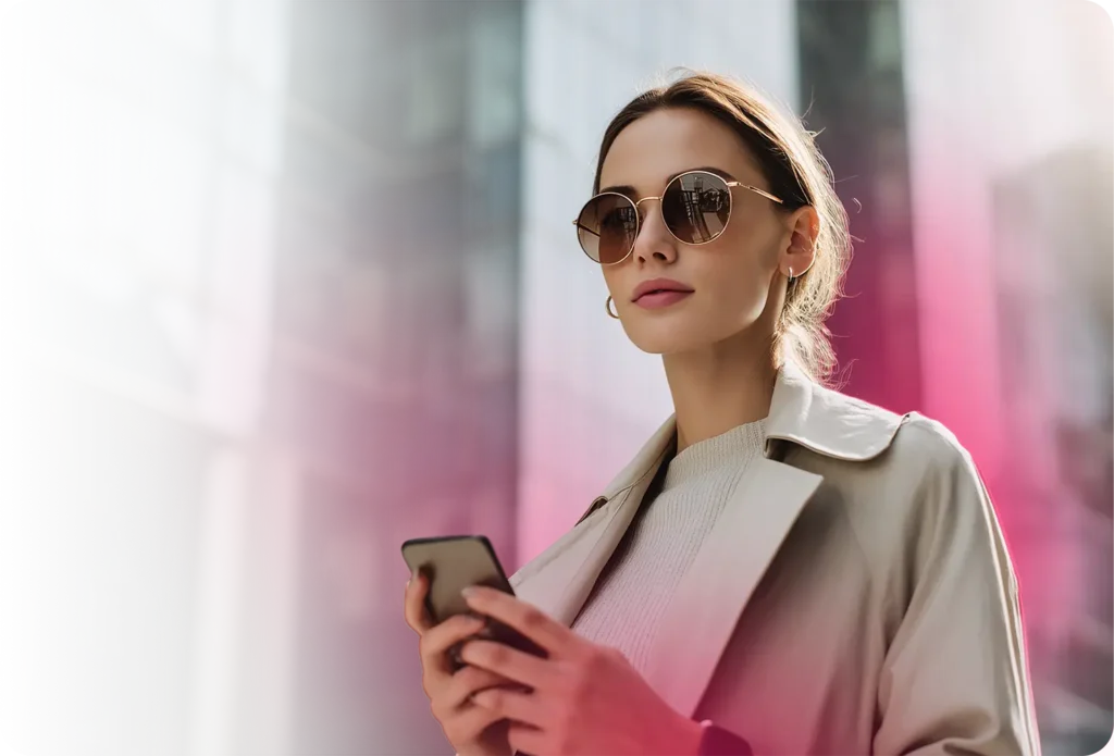 Mujer joven y elegante con gafas de sol tipo aviador y un abrigo beige sosteniendo un smartphone a su lado, mirando hacia arriba con confianza contra un horizonte urbano difuminado con un brillo bokeh rosa suave, que representa a una audiencia de consumidores móviles adinerados que viven en la ciudad