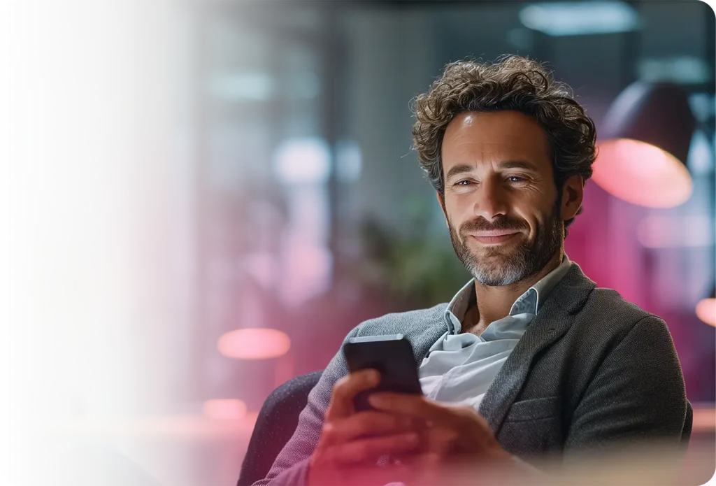 Hombre profesional seguro de sí mismo con blazer gris sosteniendo un smartphone en un salón moderno con iluminación bokeh rosa suave