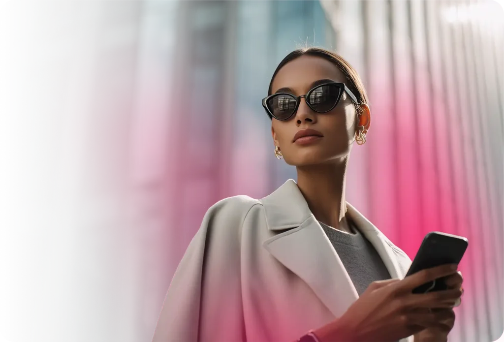Joven mujer con gafas de sol redondas de montura negra y un abrigo oversize sosteniendo un smartphone a su lado y mirando hacia arriba frente a columnas arquitectónicas suavemente difuminadas con un fondo bokeh rosa, que representa a un consumidor móvil urbano con estilo orientado hacia la moda y dirigido a través de la plataforma publicitaria de TrueSignal