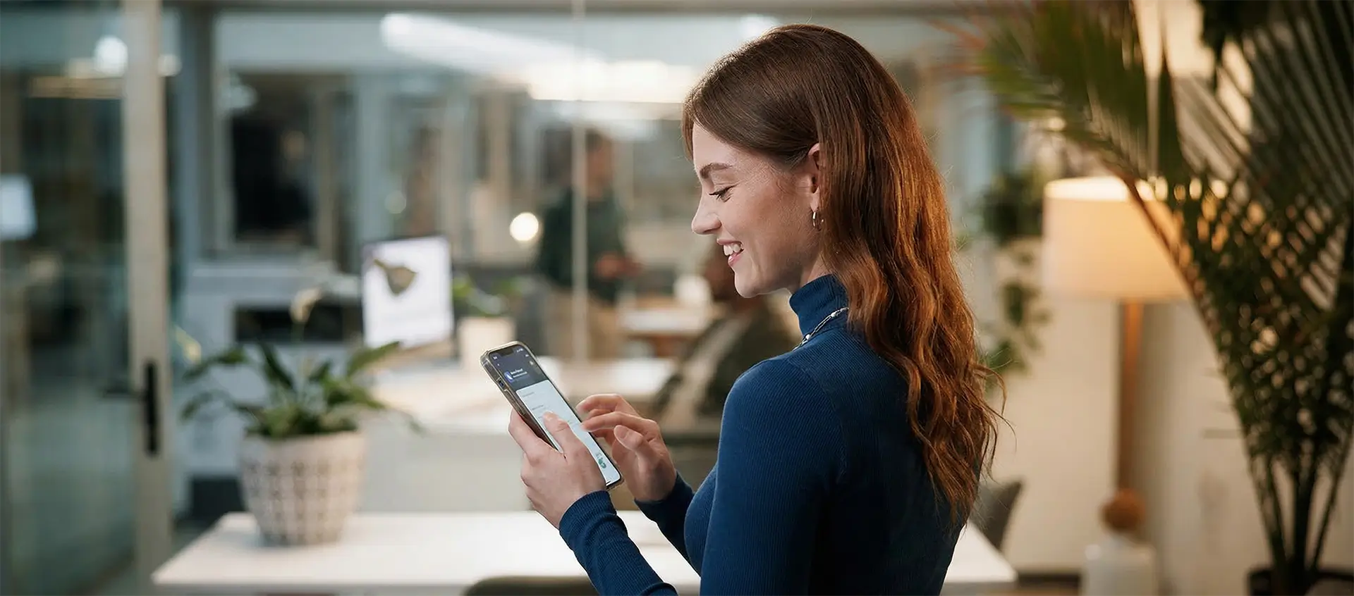 Joven profesional sonriendo mientras usa un smartphone en una oficina moderna con luz natural y plantas.